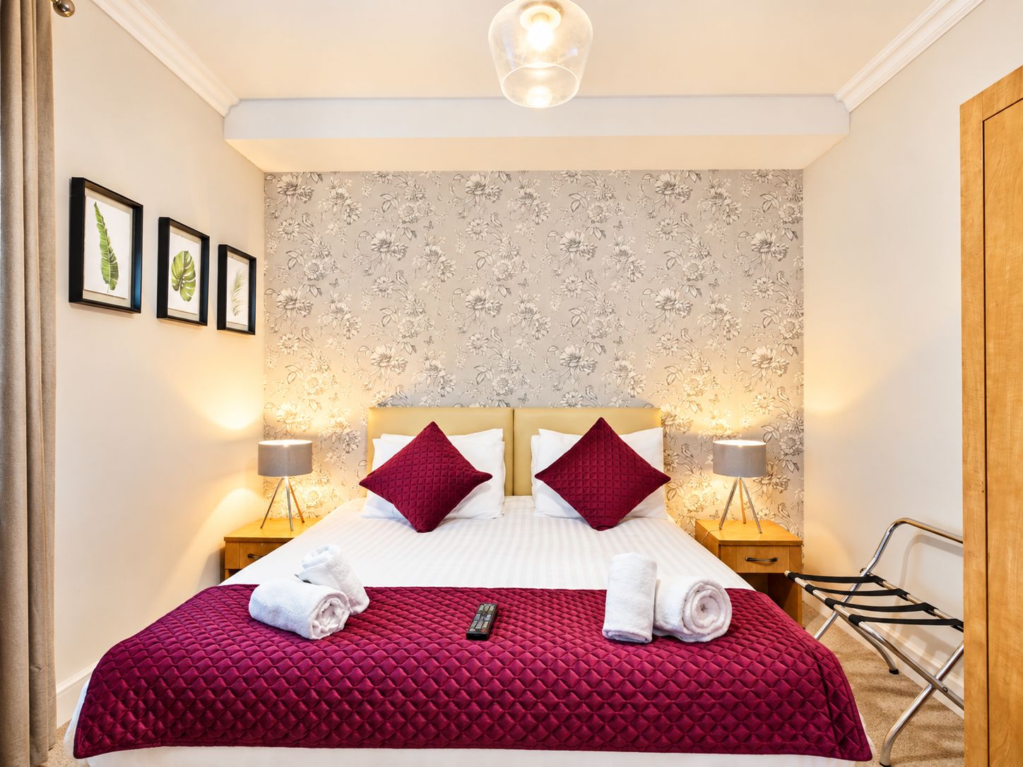 A hotel room with a bed, two bedside lamps, a wall with floral wallpaper, and framed botanical prints on the wall.