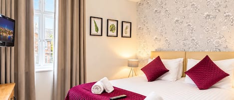 Una habitación de hotel con cama, televisor, ventana con cortinas y papel tapiz floral en la pared.