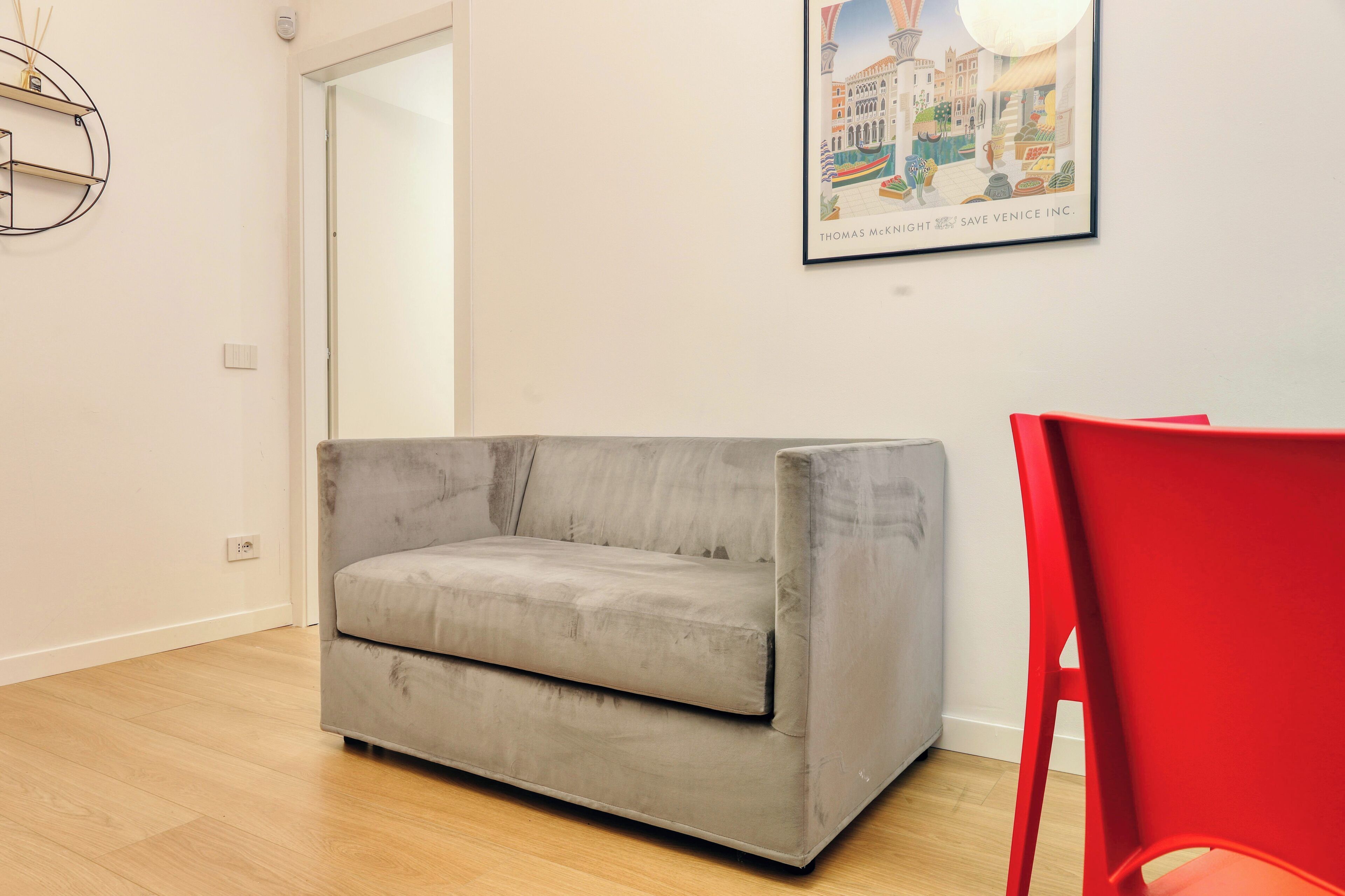 A room with a grey sofa, a red chair, and a framed picture of a cityscape on the wall.