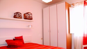 A bedroom with a bed, red bedding, a white wardrobe, and two wooden birdcages on a shelf.