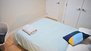 Een eenpersoonsbed met een opgevouwen witte handdoek, een kleurrijk kussen en een witte stoel in een kamer met witte muren en een houten vloer.