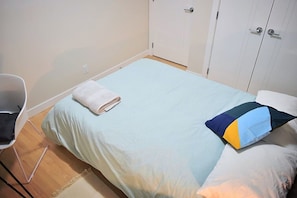 Un letto singolo con un asciugamano bianco piegato, un cuscino colorato e una sedia bianca in una stanza dalle pareti bianche e pavimento in legno.