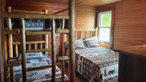 A cabin room with a bunk bed, a wooden ladder, a window with a view of greenery, and a quilted bedspread.