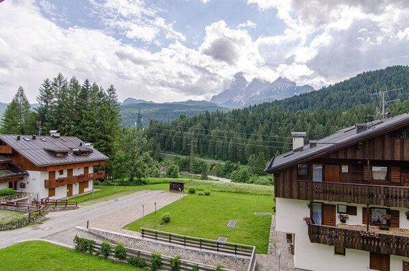 A traditional chalet-style building with a wooden facade, a balcony with flower boxes, and a view of a mountainous landscape. - Summer in the mountains (San Vito di Cadore)
