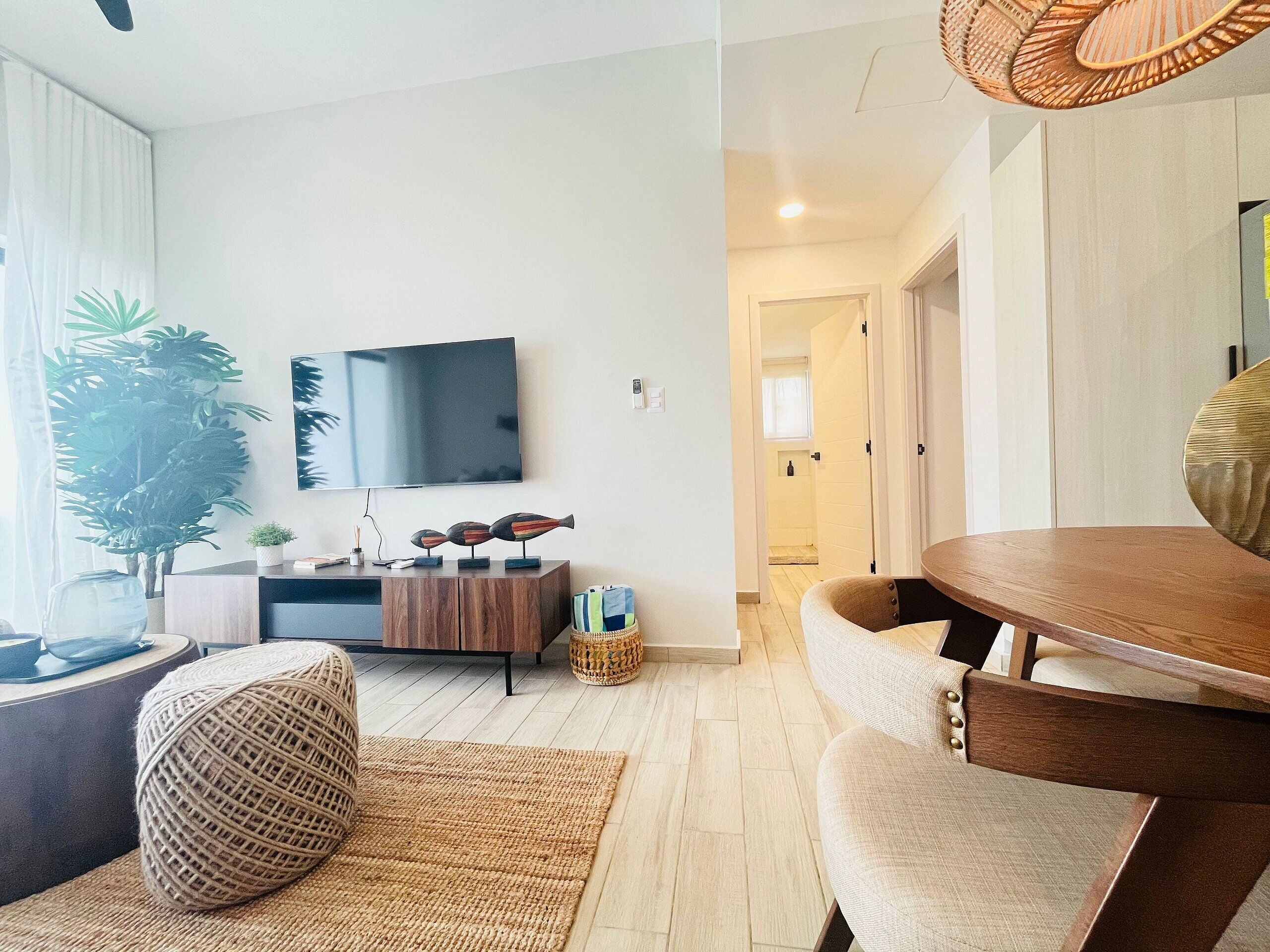 A modern living room with a flat-screen TV, a large plant, a wooden coffee table, and a cozy seating area.