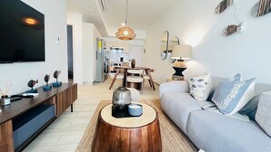 Un salon moderne avec un canapé, une table basse et un téléviseur. On aperçoit un coin repas au fond de la pièce.
