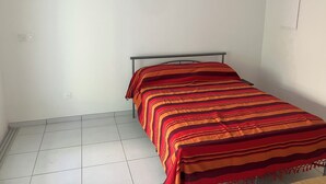 A small, single-bed room with a metal frame bed, white walls, and a tiled floor. - Studio 5 minutes from Sainte Anne beaches and hiking trails (Sainte-Anne)