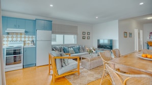 A modern kitchen with blue cabinets, a white refrigerator, and a stove. There is a dining area with a table, chairs, and a TV.
