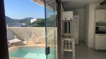 Varanda envidraçada com piscina, mobiliário branco e área de cozinha.
