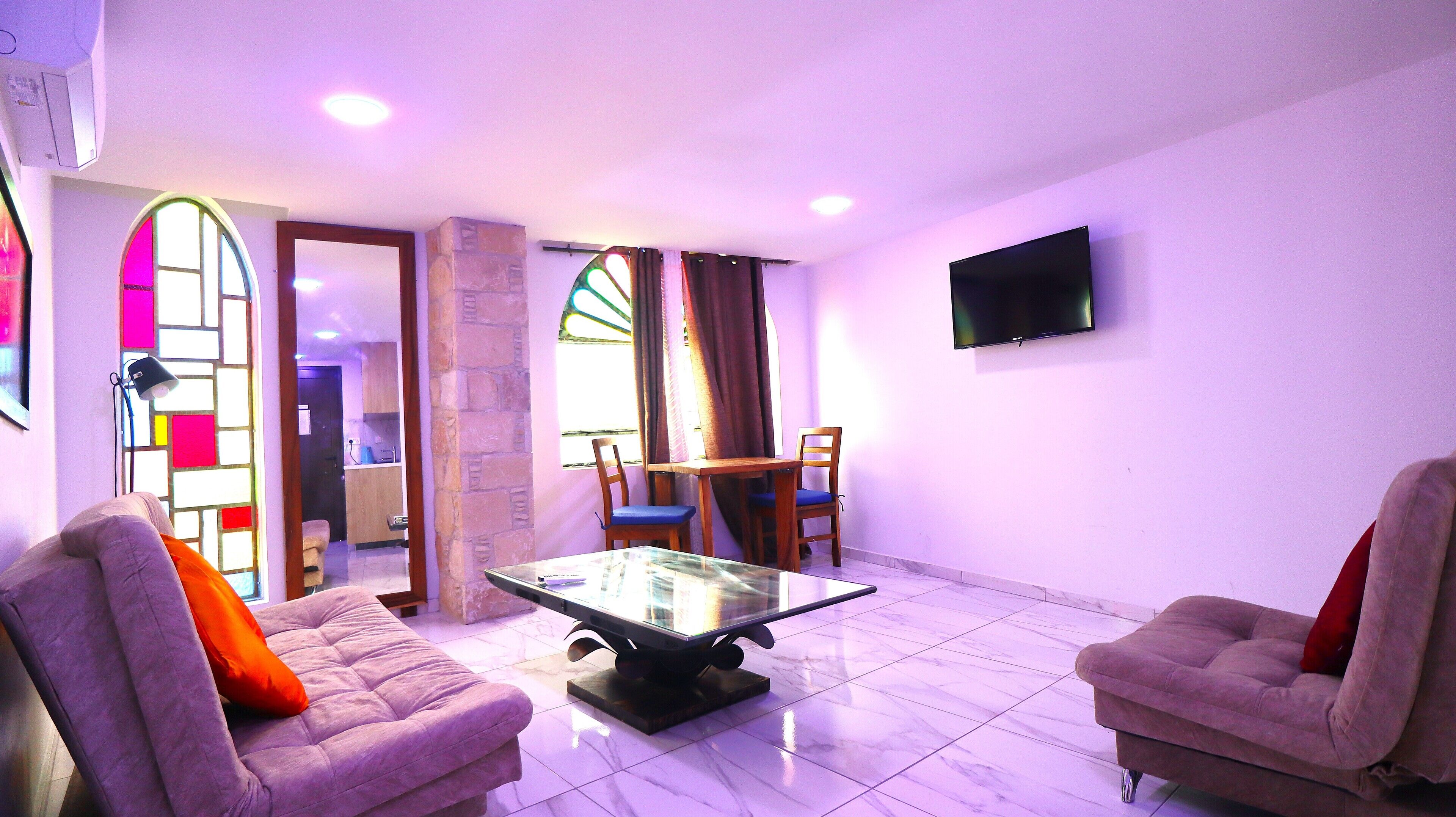 A living room with two sofas, a glass coffee table, a TV mounted on the wall, and a dining area with a table and chairs.