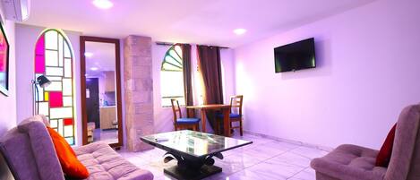 Una sala de estar con dos sofás, una mesa de centro de vidrio, un televisor montado en la pared y una zona de comedor con mesa y sillas.