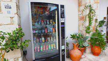 Máquina expendedora con diversas bebidas y aperitivos, un código QR para WiFi y plantas en macetas cerca.