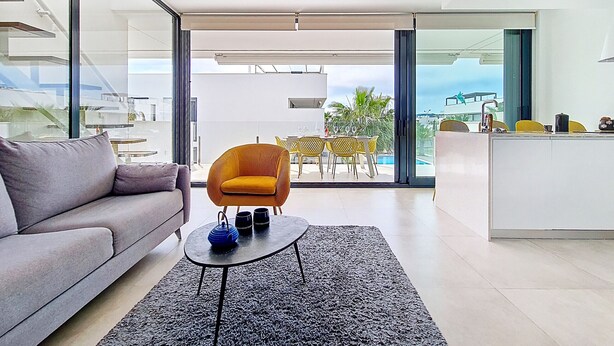 A modern living room with a sofa, a coffee table, and a chair. There is a view of a pool and outdoor furniture through large glass doors. - Penthouse apartment with pool views (Mar de Cristal)