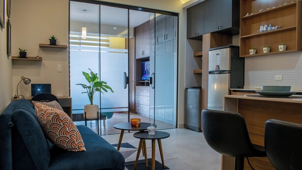 A modern living room with a sofa, coffee table, and chairs. There is a sliding glass door leading to a kitchen area with a refrigerator and cabinets.
