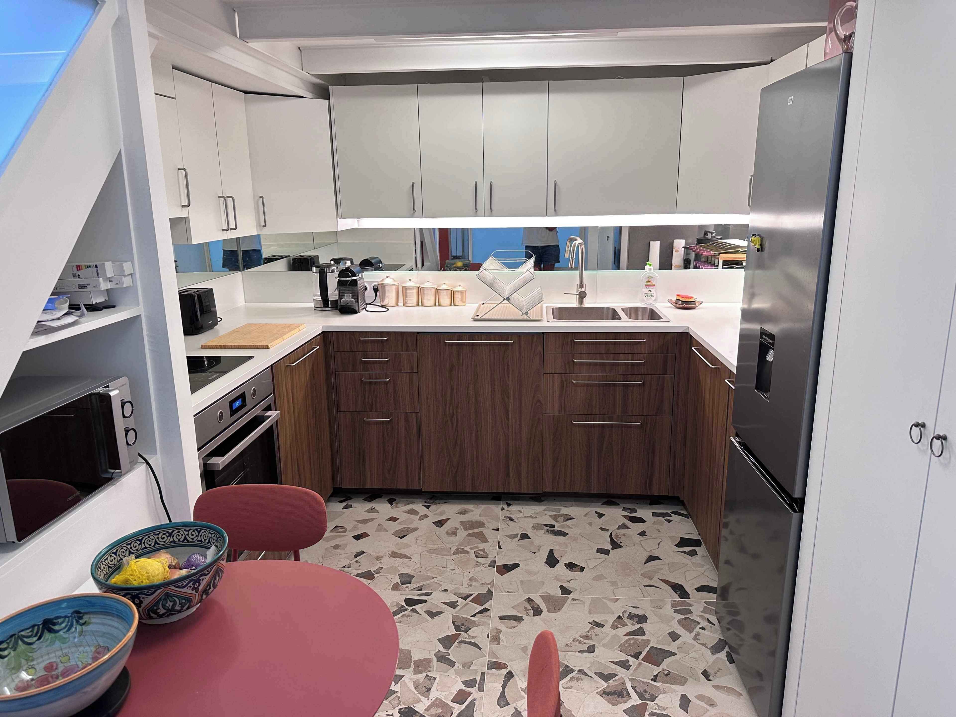 A compact kitchen with white cabinets, a stainless steel refrigerator, a built-in oven, and a small dining area with a red table and two bowls.