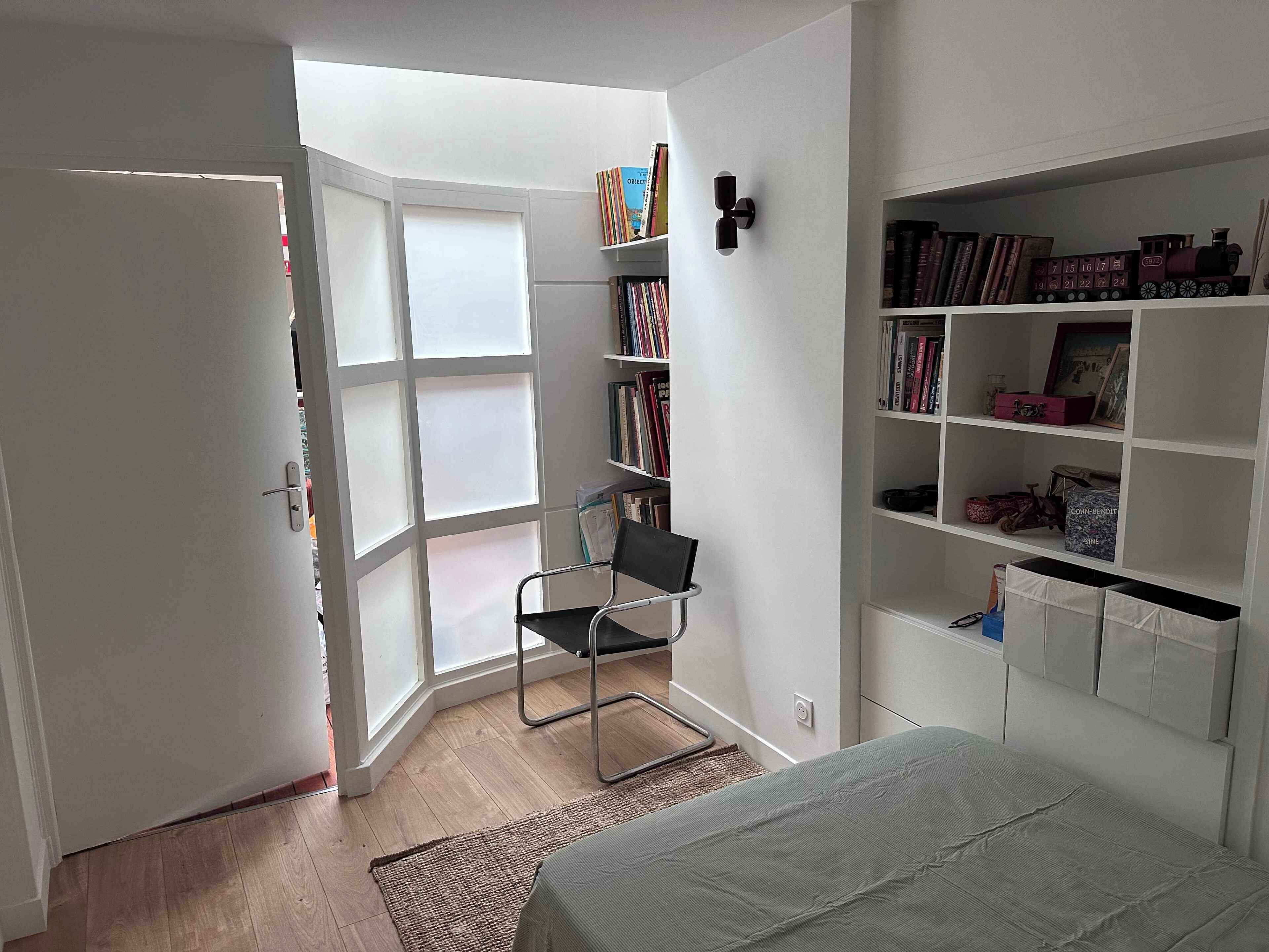A room with a bed, a chair, a bookshelf, and a window.