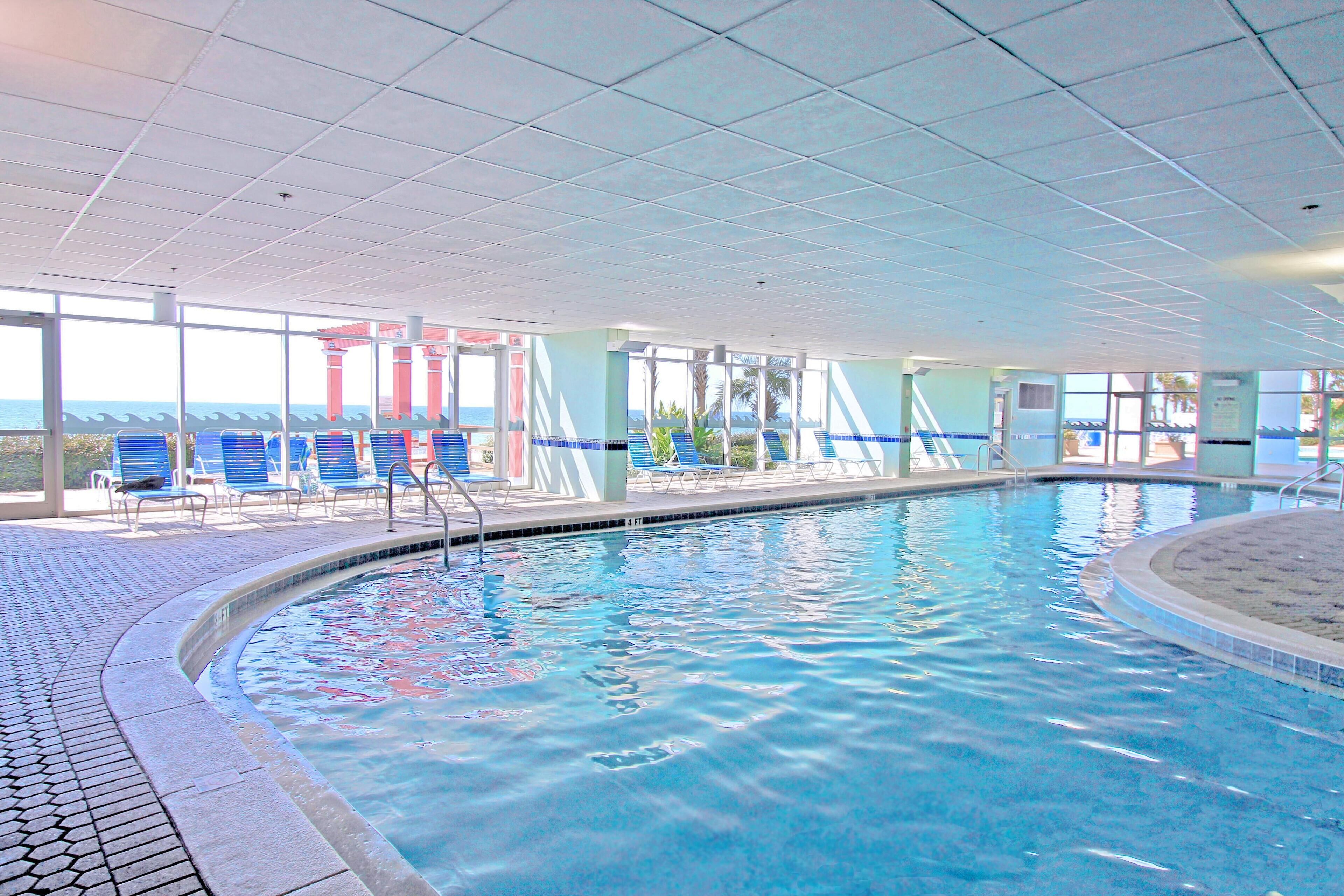 Una piscina interior con agua azul cristalina, rodeada de sillas reclinables azules y un suelo de baldosas.