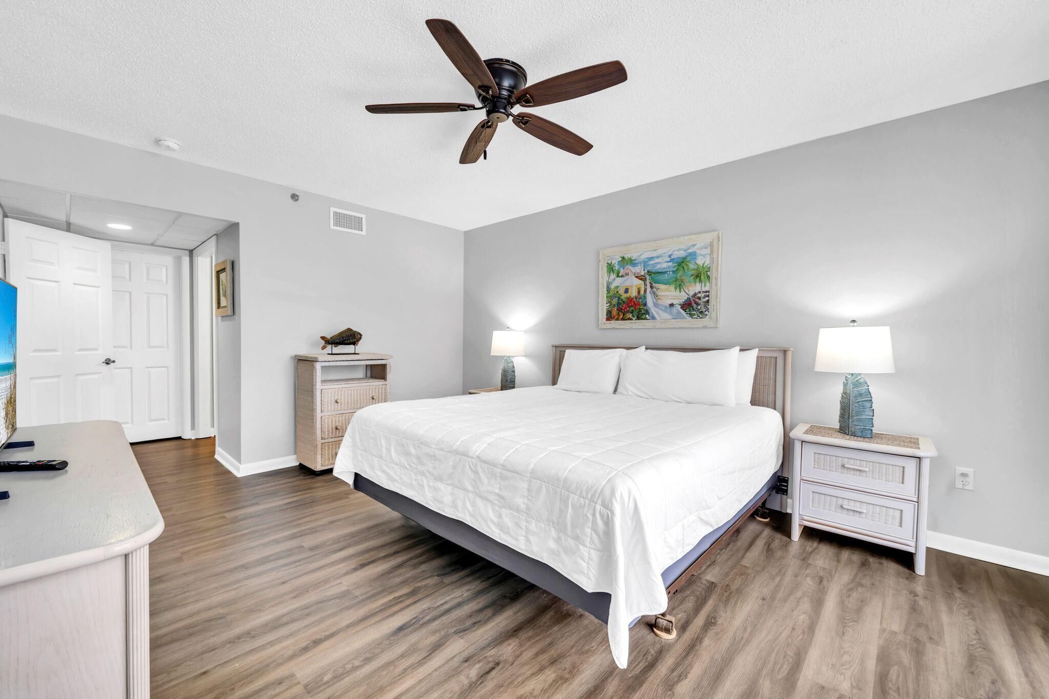 A bedroom with a large bed, a ceiling fan, a nightstand with a lamp, a dresser, and a painting on the wall.
