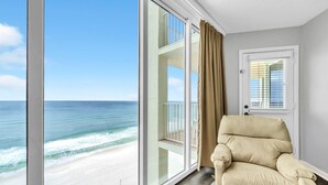 Una habitación con un ventanal amplio que da a la playa, un sillón color crema y una puerta con persianas.