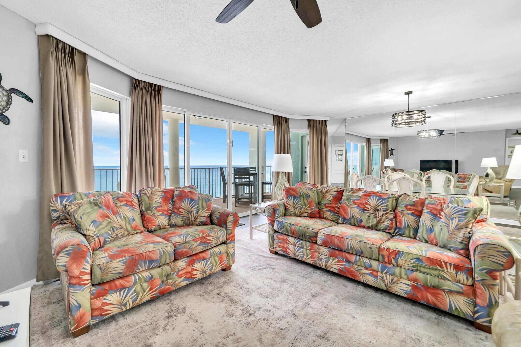 A spacious living room with two floral-patterned sofas, a ceiling fan, and a dining area with white chairs.
