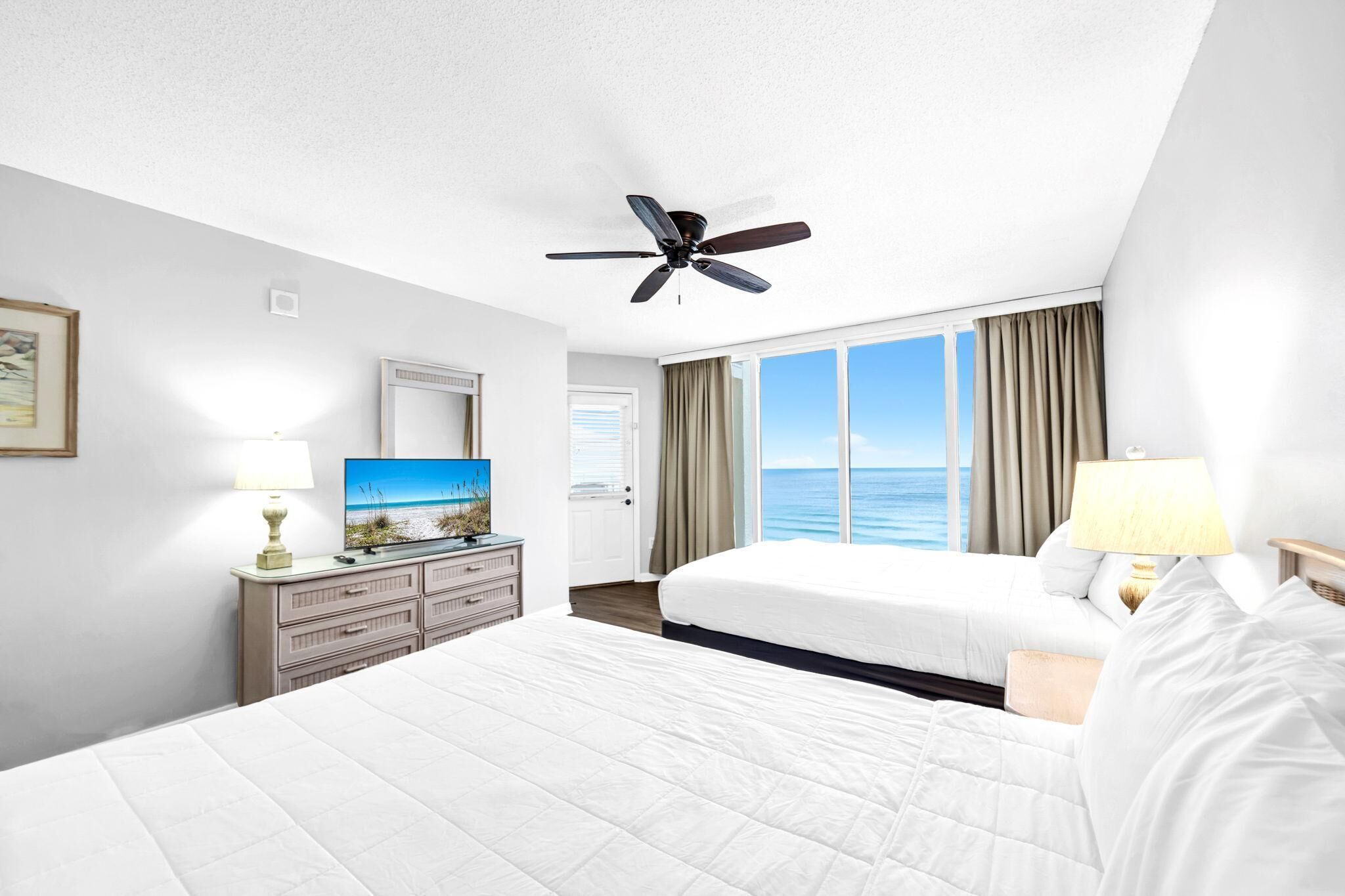 A hotel room with a large bed, a ceiling fan, a TV, and a view of the ocean.