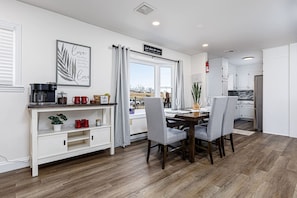 A modern kitchen with white cabinets, a dining area with a wooden table and grey chairs, and a coffee station with mugs and a coffee maker.