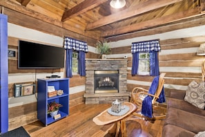 A log cabin interior with a stone fireplace, wooden walls, a blue bookshelf, a television, and a rocking chair.