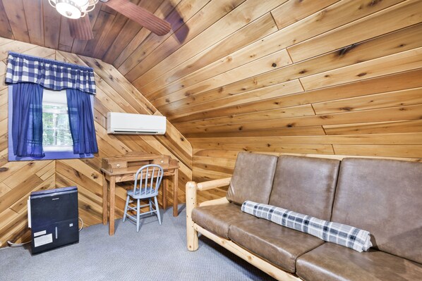 Una habitación acogedora tipo cabaña, con paredes de madera, un sofá de cuero marrón, una manta escocesa, un escritorio con silla y un equipo de aire acondicionado.