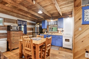 Interior rústico de una cabaña con una mesa de comedor de madera, armarios azules, una estufa blanca y techo de madera.