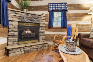 A log cabin interior with a stone fireplace, wooden furniture, and a window with checkered curtains. - Woodland view-Blue Log Cabin (Laurelville)