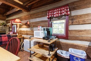 A rustic cabin interior with a wooden dining table, a red chair, a microwave, a coffee maker, and a window with red curtains.