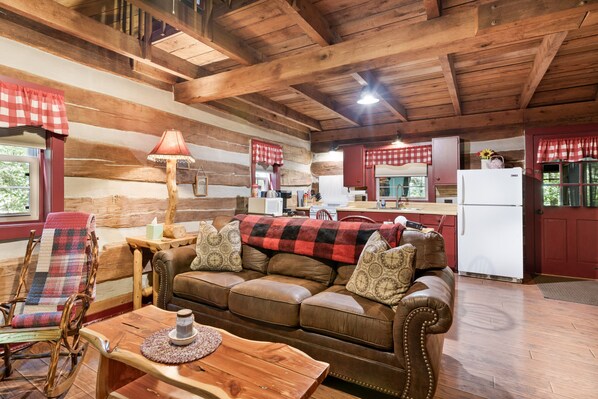 A cozy cabin interior with a wooden ceiling, a leather sofa with patterned cushions, a rocking chair, a wooden table, and a kitchen area with a refrigerator and cabinets.