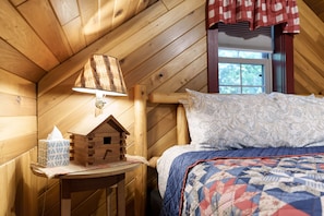 Ein holzgetäfelter Raum im Blockhausstil mit einem Bett, einem Nachttisch mit Lampe und einem Fenster mit Karottenmuster-Vorhang.