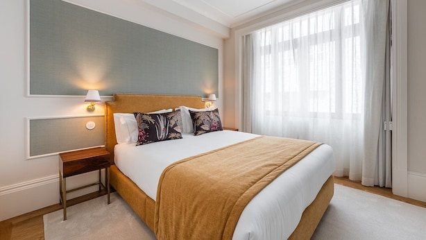 Una habitación de hotel con una cama grande, una mesita de noche, un televisor montado en la pared y una ventana con cortinas translúcidas.