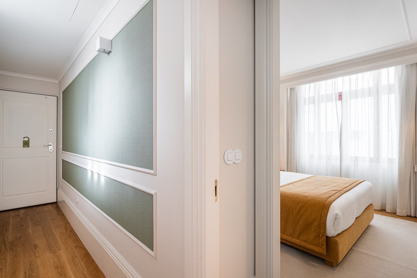 A hotel room with a bed, a door with frosted glass panels, and a view of the outside through sheer curtains.