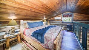 Une chambre confortable dans un chalet, avec un plafond en bois, un lit recouvert d’une literie à motifs, une table de chevet avec une lampe, et une vue sur l’extérieur par une fenêtre.