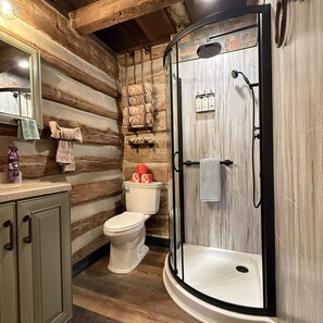 A bathroom with a wooden log wall, a toilet, a shower with a glass door, and a sink with a mirror. - Woodland view-Green Log Cabin (Laurelville)