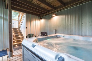 A hot tub in a rustic cabin with wooden stairs leading to a door, a chair, and a metal heater on the wall.