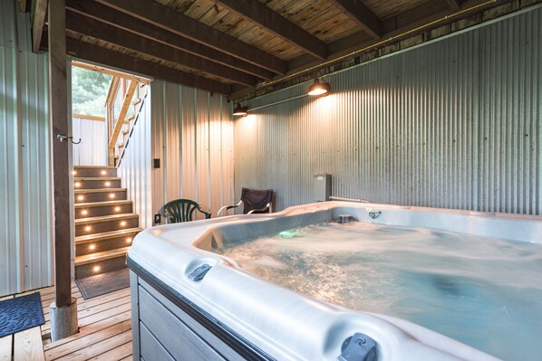 A hot tub in a rustic cabin with wooden stairs leading to a door, a chair, and a metal heater on the wall. - Woodland view-Hitchin' Post (Laurelville)