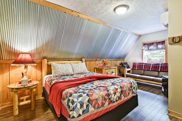 A cozy bedroom with a wooden headboard, a quilted bedspread, a small bedside table with a lamp, and a bench with a plaid blanket.