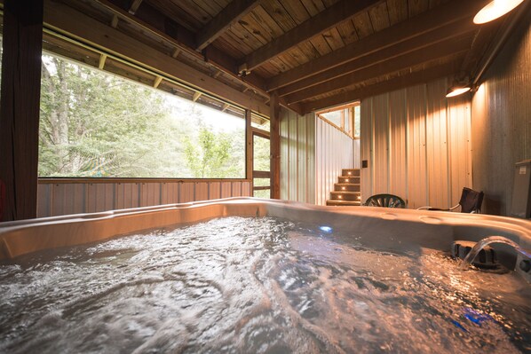 A hot tub with a wooden interior, a window with a view of trees, and a chair near the window.