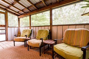 A covered porch with wooden flooring, two upholstered chairs, and large windows offering a view of trees.