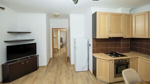 A compact kitchen with a refrigerator, stove, and wooden cabinets. A flat-screen TV is mounted on a dark cabinet.