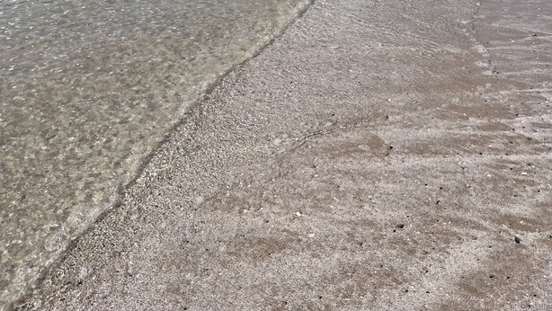 Ein Strand mit klarem Wasser, kiesigem Ufer und einer fernen Küstenlinie.