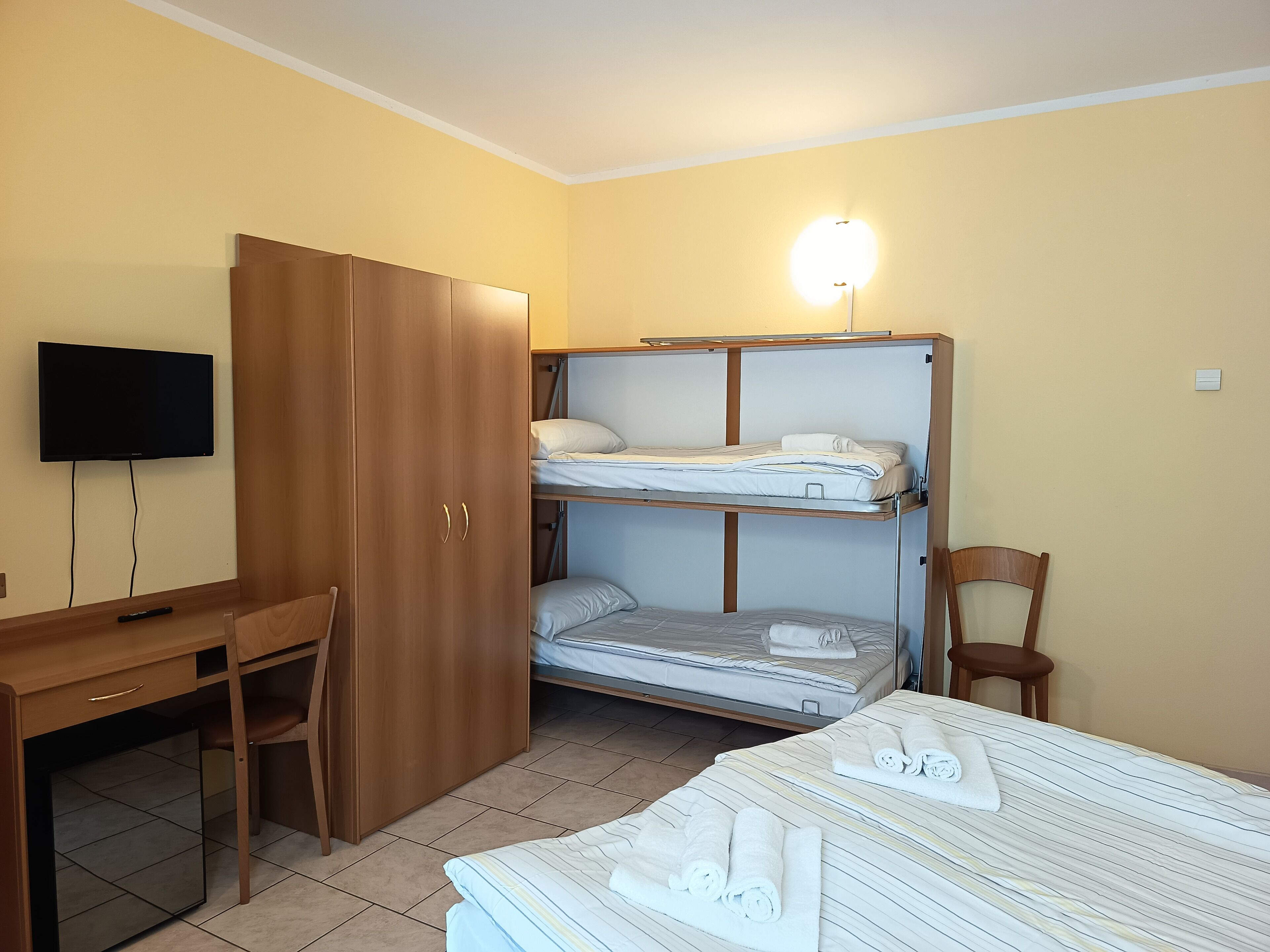 A room with a bunk bed, a desk with a chair, a television, and a bedside table with towels.