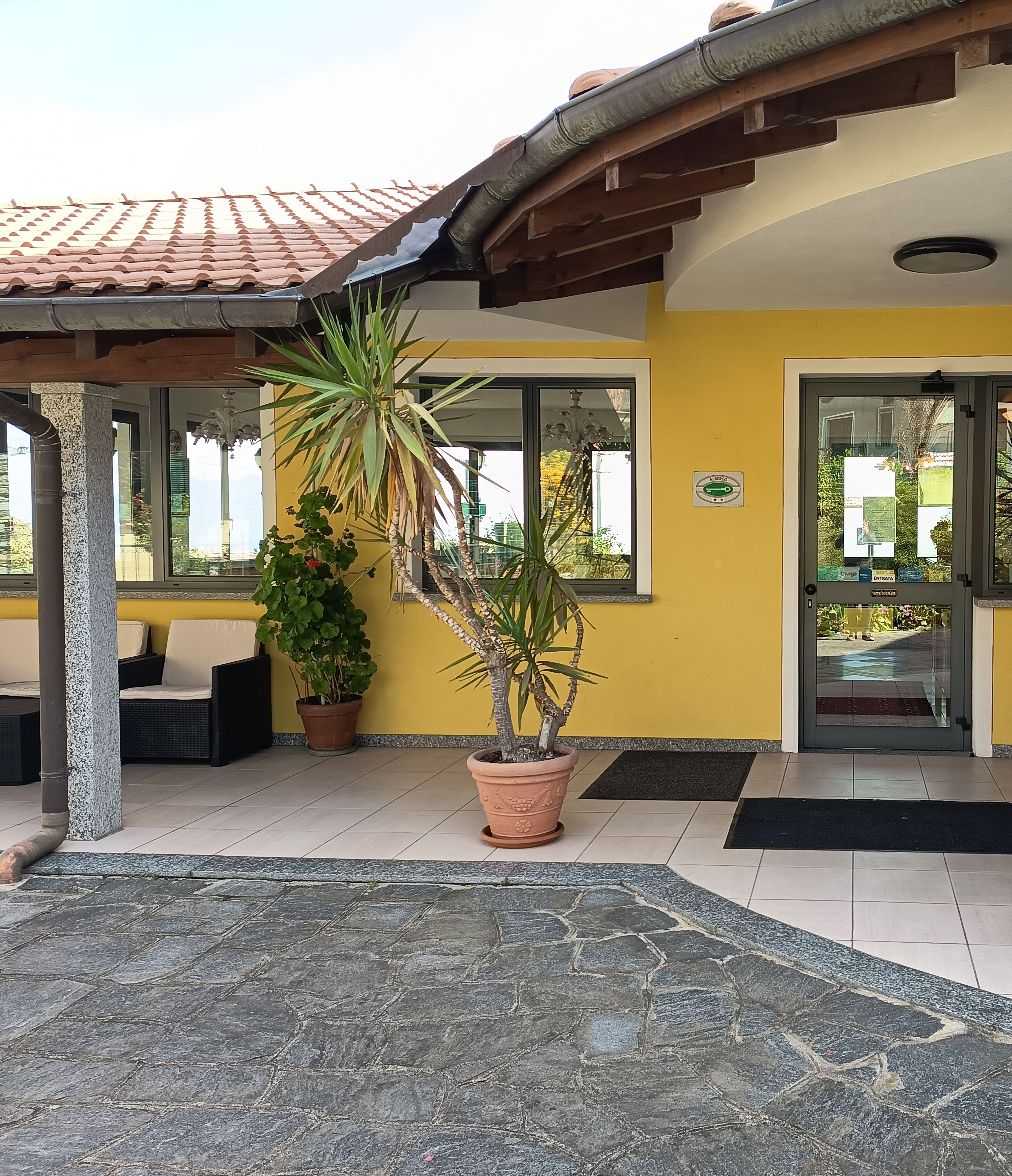 A building entrance with a tiled roof, a potted plant, a bench, and a door with a glass panel.