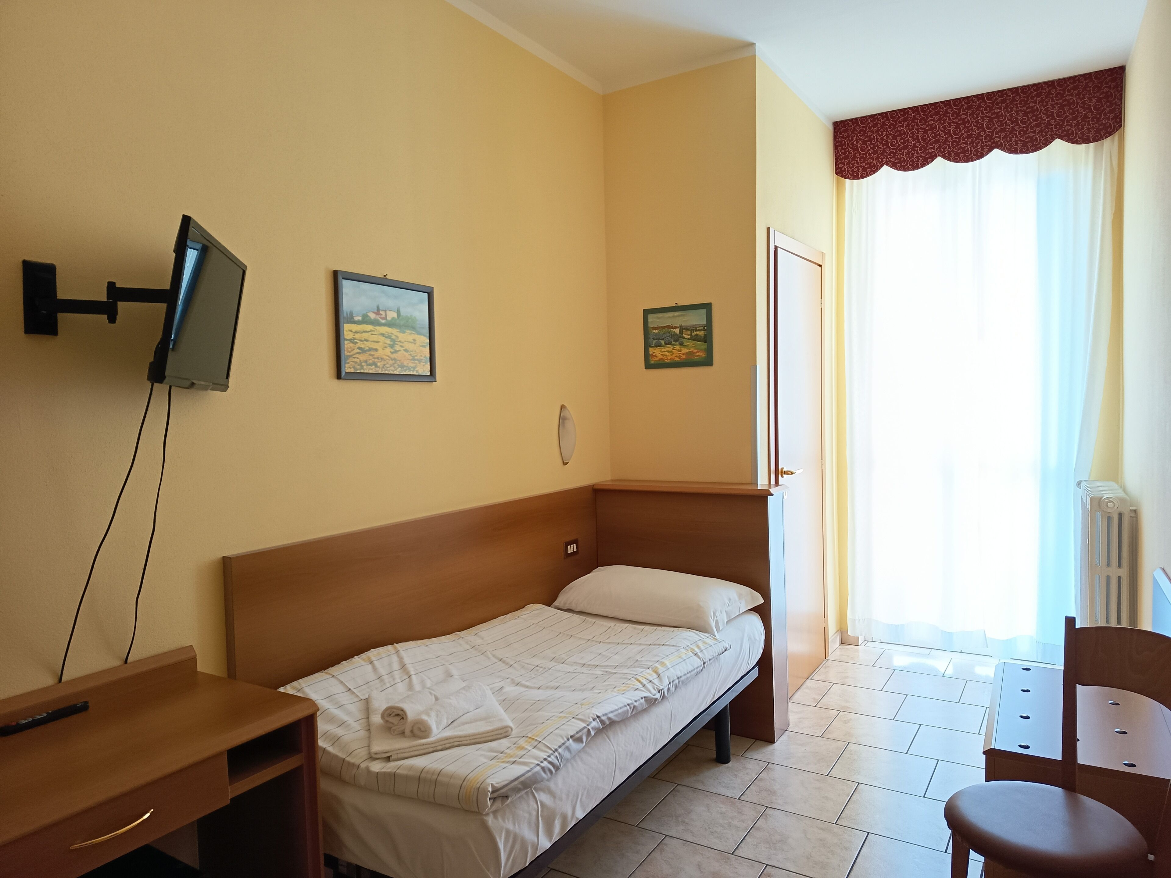 A single bed with a wooden headboard, a bedside table with a lamp, a TV mounted on the wall, and two paintings on the wall.