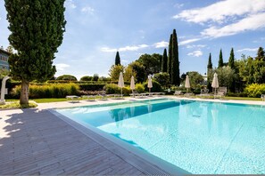 A rectangular swimming pool with a tiled deck, surrounded by lounge chairs and umbrellas, with tall trees and a clear sky in the background. - Corte Fasanella 50 in Toscolano Maderno (Toscolano Maderno)