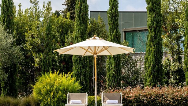 A swimming pool with two lounge chairs and a sun umbrella, surrounded by tall trees and a modern building in the background. - Corte Fasanella 50 in Toscolano Maderno (Toscolano Maderno)