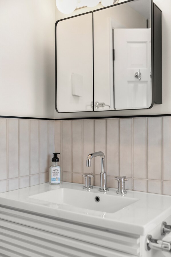 Un baño con un lavamanos blanco, un espejo encima y una botella de jabón para manos sobre el mostrador.